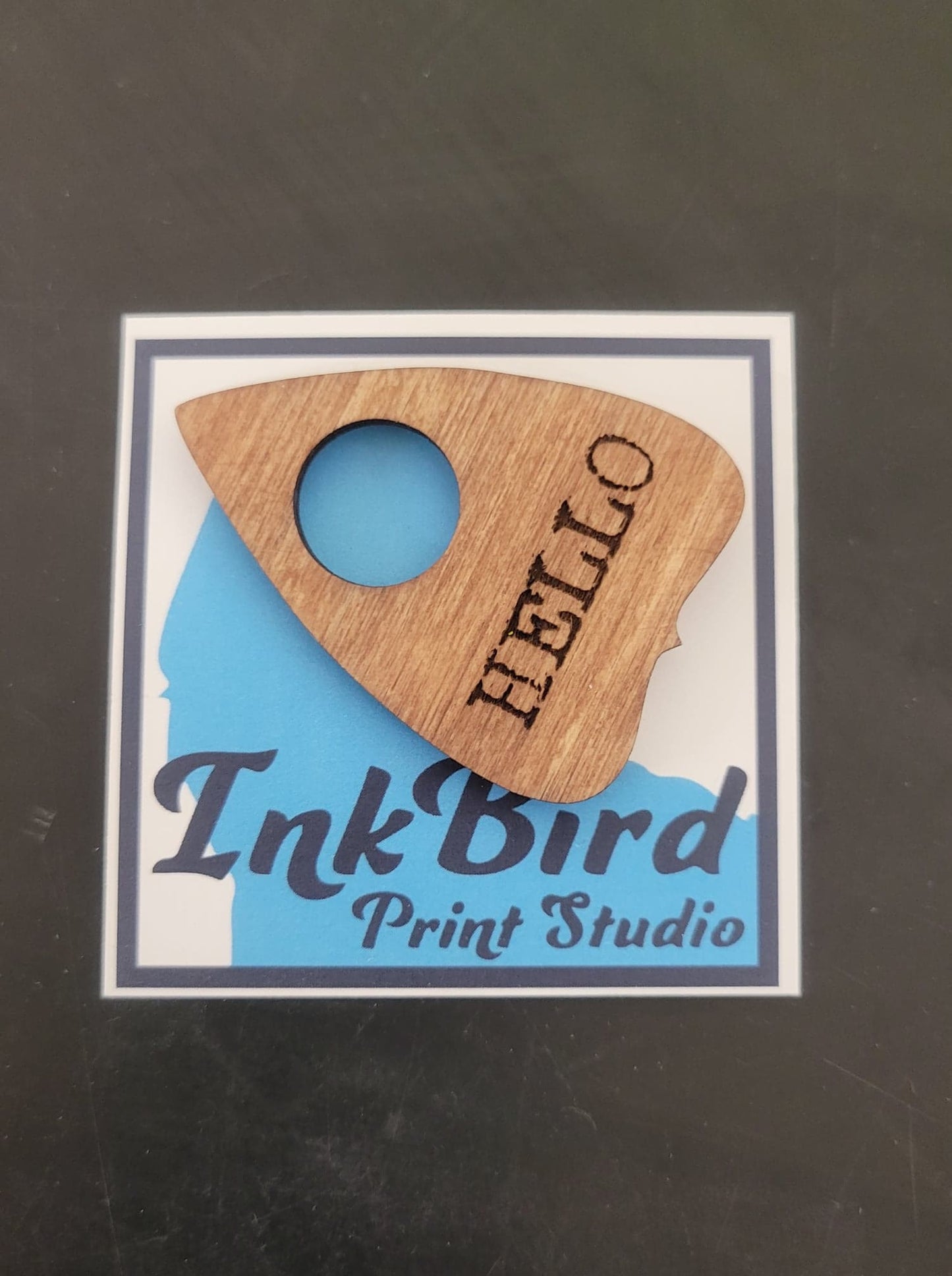 Laser-cut wooden planchette pin by InkBird Print Studio, engraved “HELLO,” available in large or small, displayed on branded backing card.