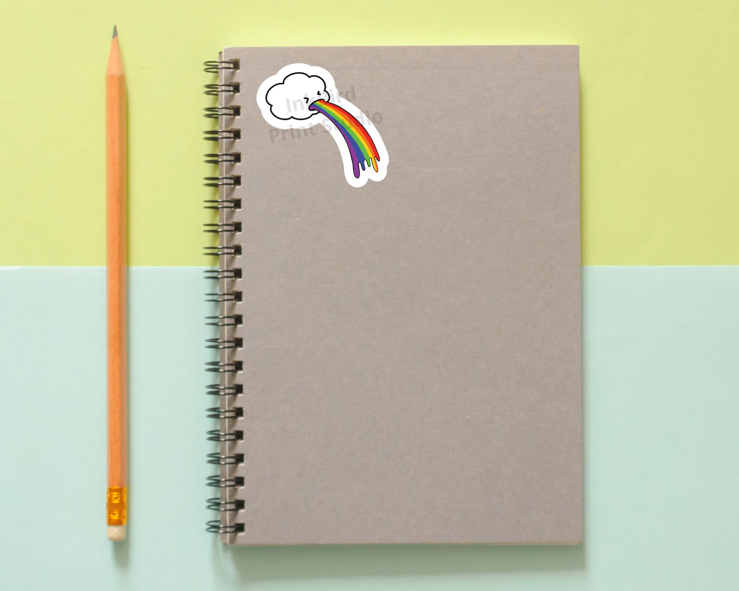 Brown spiral notebook with a glossy vinyl Rainbow Cloud Sticker by InkBird Print Studio, next to a pencil on pastel yellow and blue.