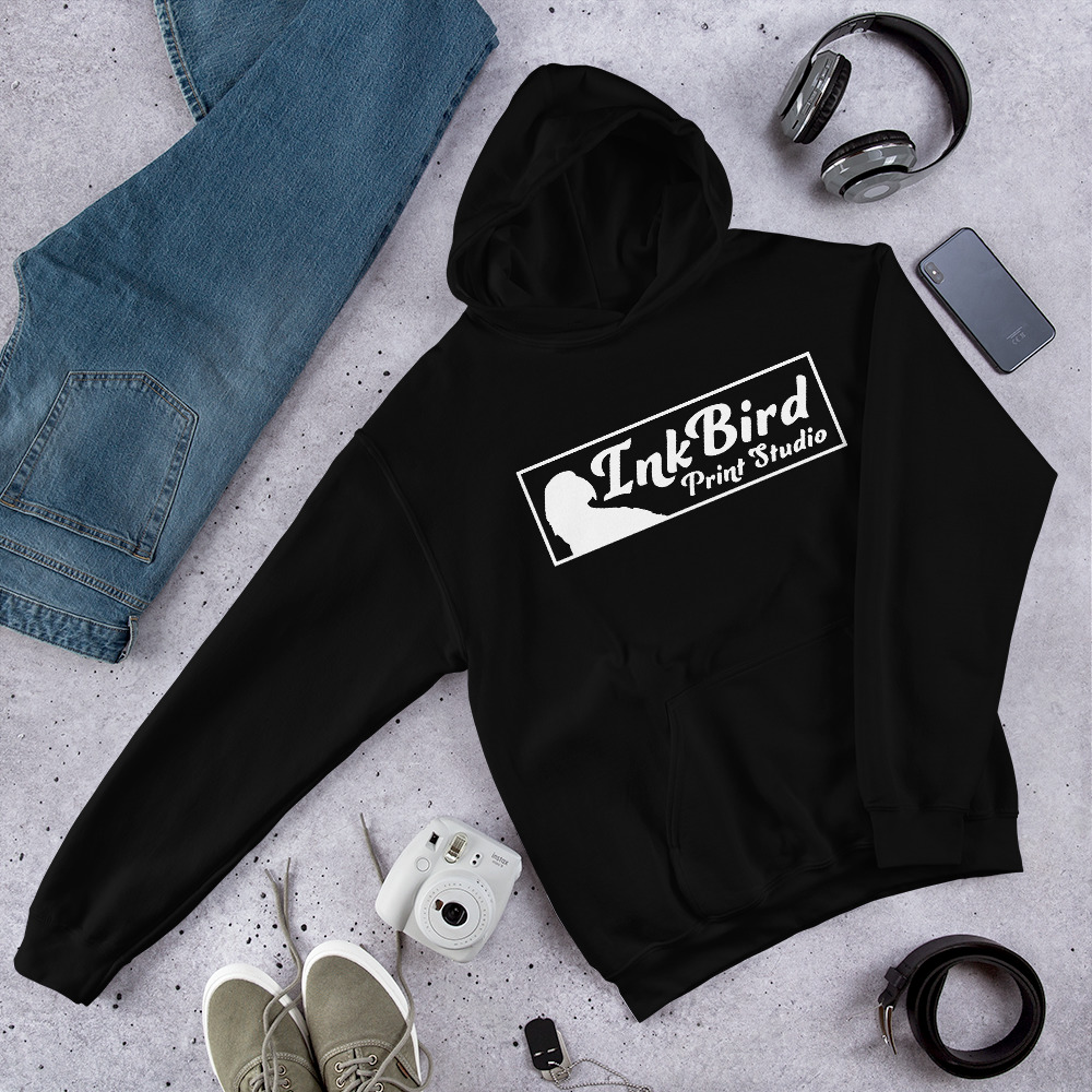 Black hoodie with “InkBird Print Studio” logo laid flat beside blue jeans, green shoes, headphones, a smartphone, instant camera, watch, and a plant on a light gray surface.