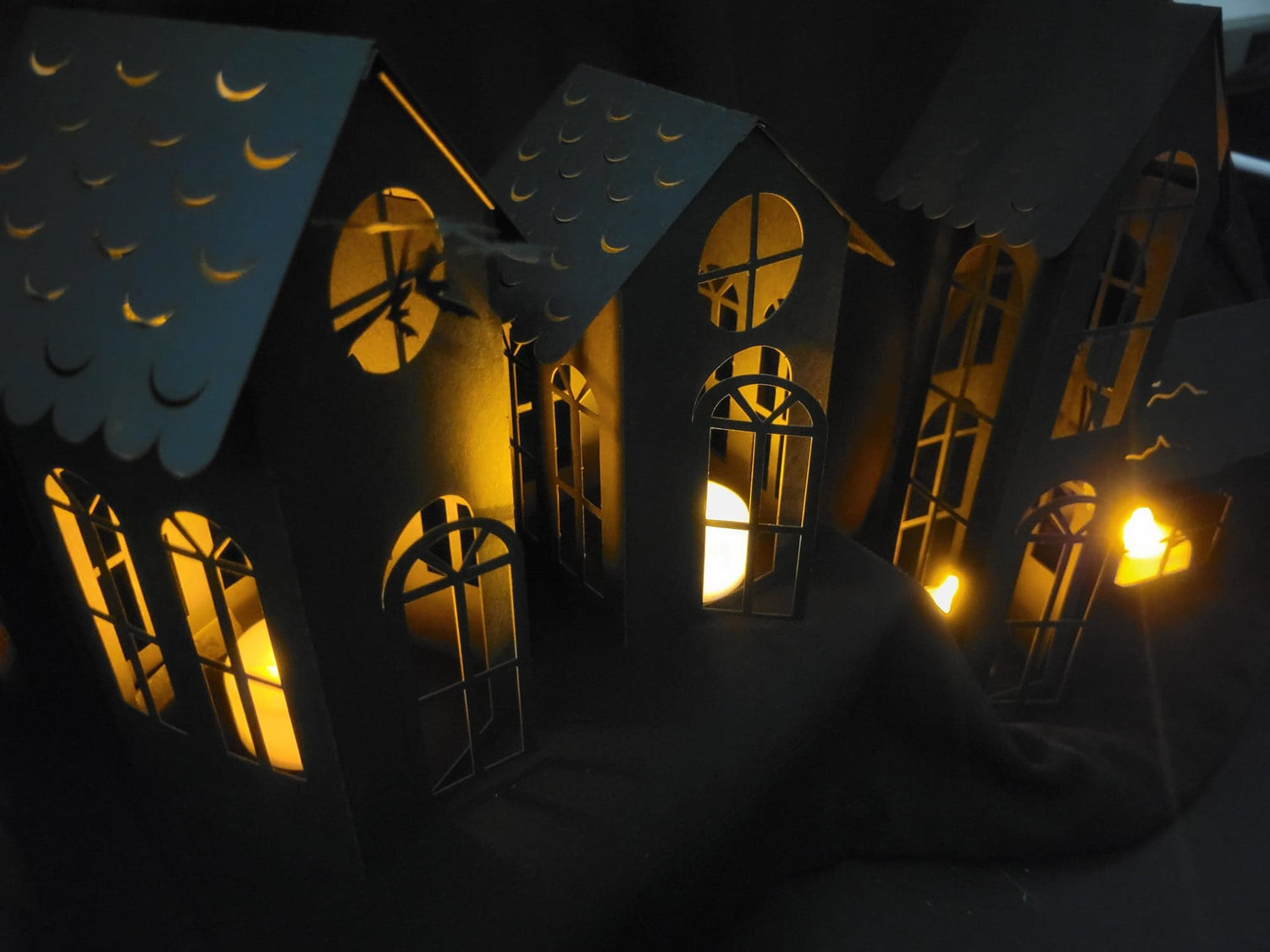Three black paper lanterns shaped like haunted houses glow with warm yellow lights inside, creating a spooky and cozy Halloween atmosphere against a dark background.