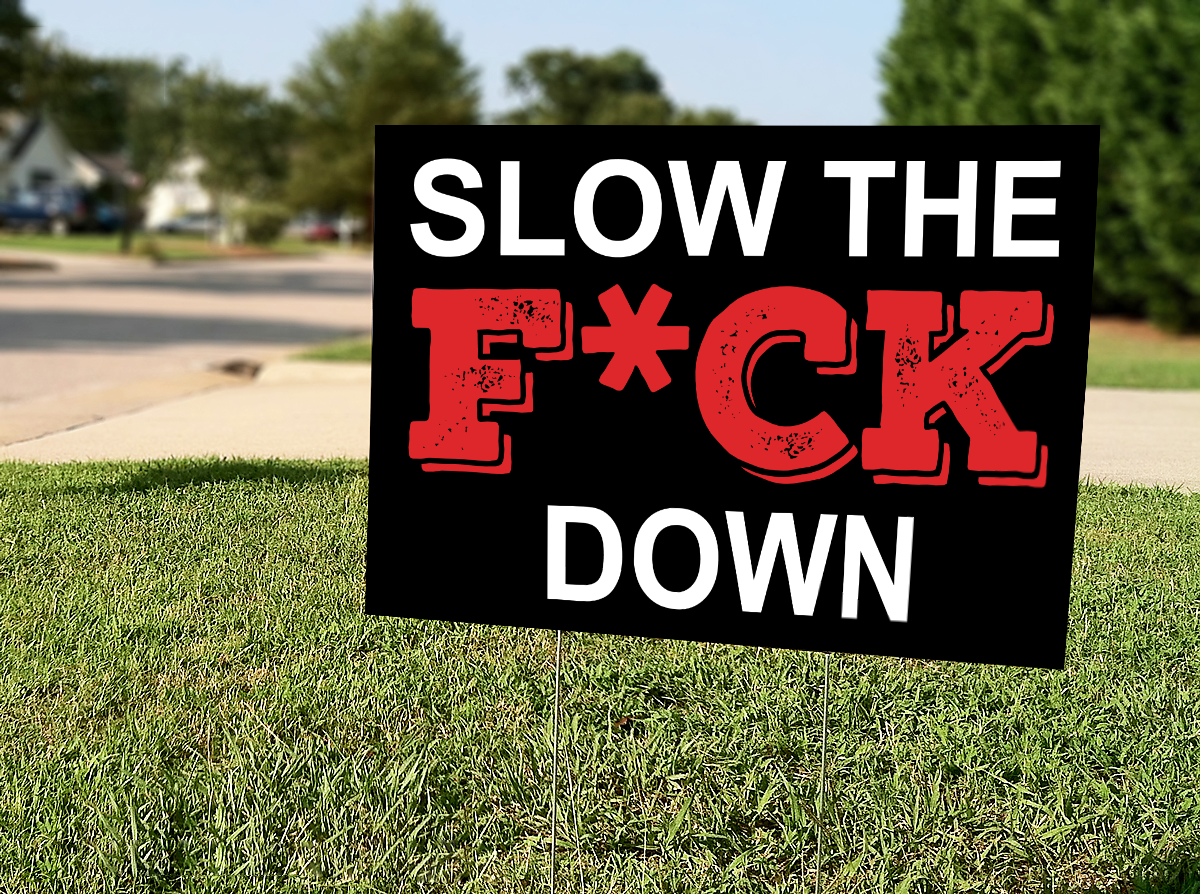 A black yard sign on grass by a street reads SLOW THE F*CK DOWN in bold white and red letters, with asterisk censoring the expletive.