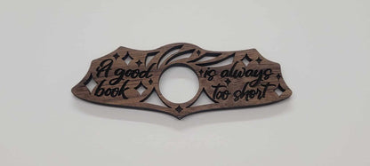 The Laser Cut Wood Book Holder from InkBird Print Studio LLC is crafted from walnut wood into intricate patterns, featuring the handwritten quote, A good book is always too short. Its an ideal thoughtful gift for any book lover.