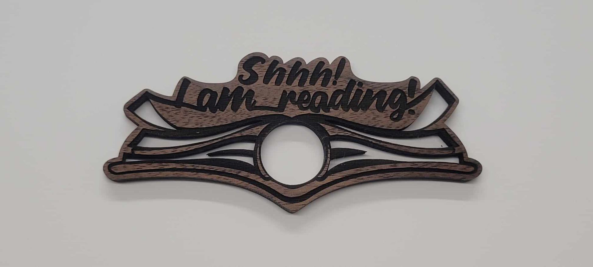 Introducing the Laser Cut Wood Book Holder by InkBird Print Studio LLC—a beautifully crafted wooden accessory perfect for book lovers. Featuring the text Shhh... I am reading! carved into exquisite Walnut wood, this design boasts two open books along with a large circle at the bottom center. The thoughtful gift highlights light brown tones accented with dark brown borders on a white background.