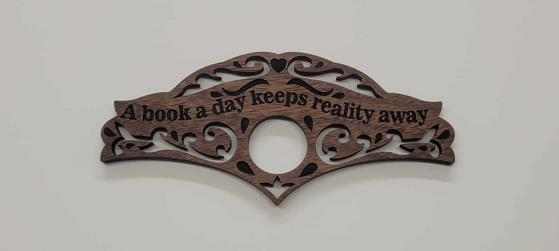 Walnut wood book holder with ornate scrollwork and A book a day keeps reality away engraved, on plain white background.