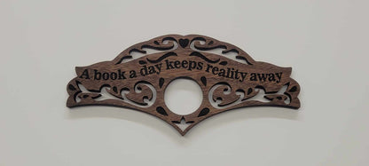 Walnut wood book holder with ornate scrollwork and A book a day keeps reality away engraved, on plain white background.