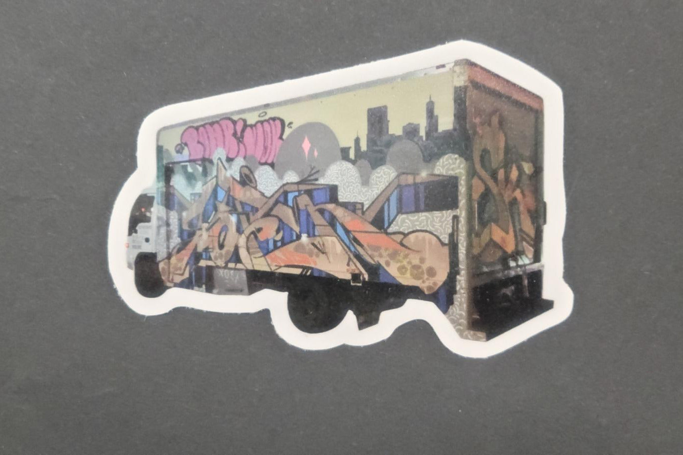 Vinyl die cut sticker of a box truck with vivid graffiti art, bright abstract shapes, and skyline on matte dark gray; strong adhesive.