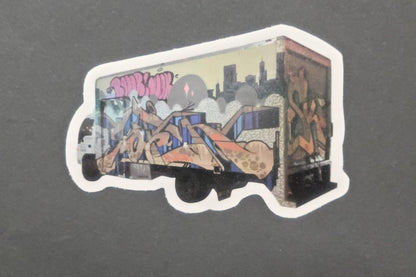 Vinyl die cut sticker of a box truck with vivid graffiti art, bright abstract shapes, and skyline on matte dark gray; strong adhesive.