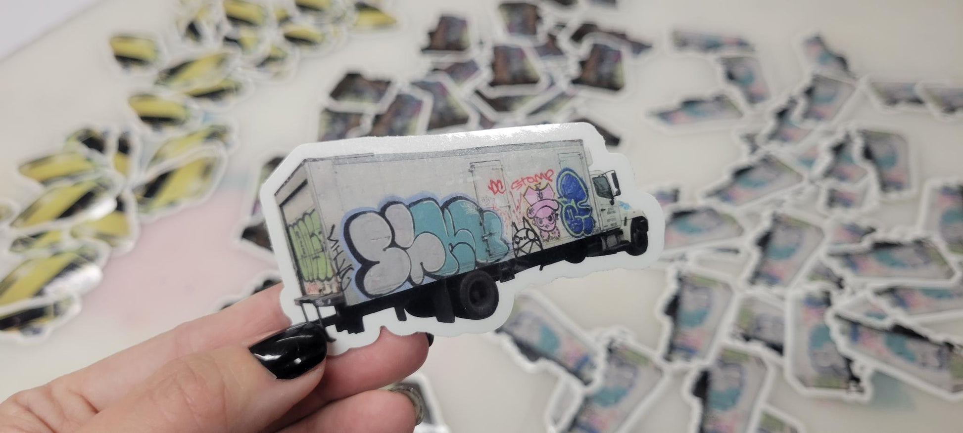 Hand with black nail polish holds a vibrant vinyl die cut sticker of a graffiti delivery truck; more colorful stickers behind.