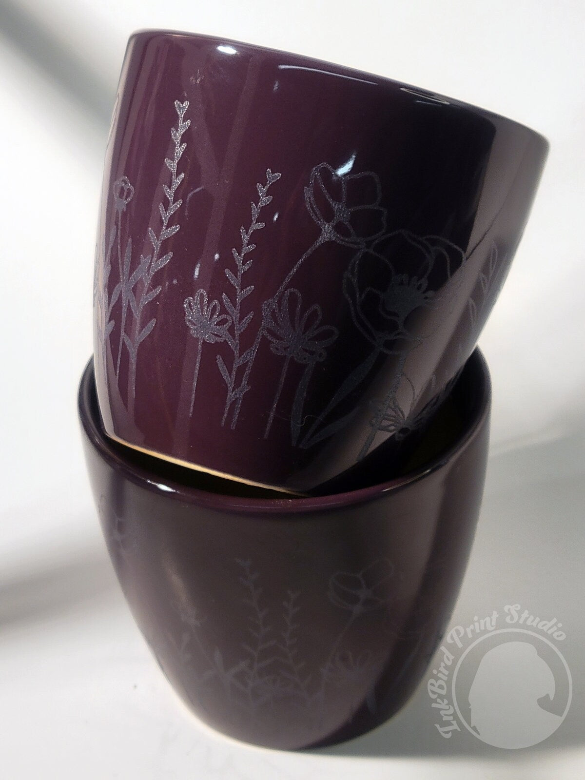 Two stacked purple ceramic mugs feature intricate botanical engravings, with a plain background accentuating the detailed floral designs on their surfaces. The bottom right corner showcases the logo of InkBird Print Studio LLC, infusing the scene with an artisanal touch.