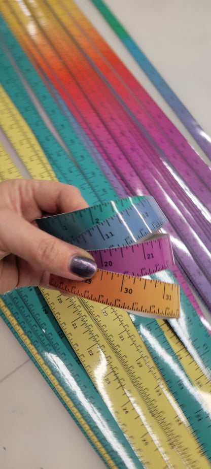 A hand holds four InkBird Print Studio adhesive vinyl rulers, showing dual inch/cm measurements, above colorful assorted rulers.