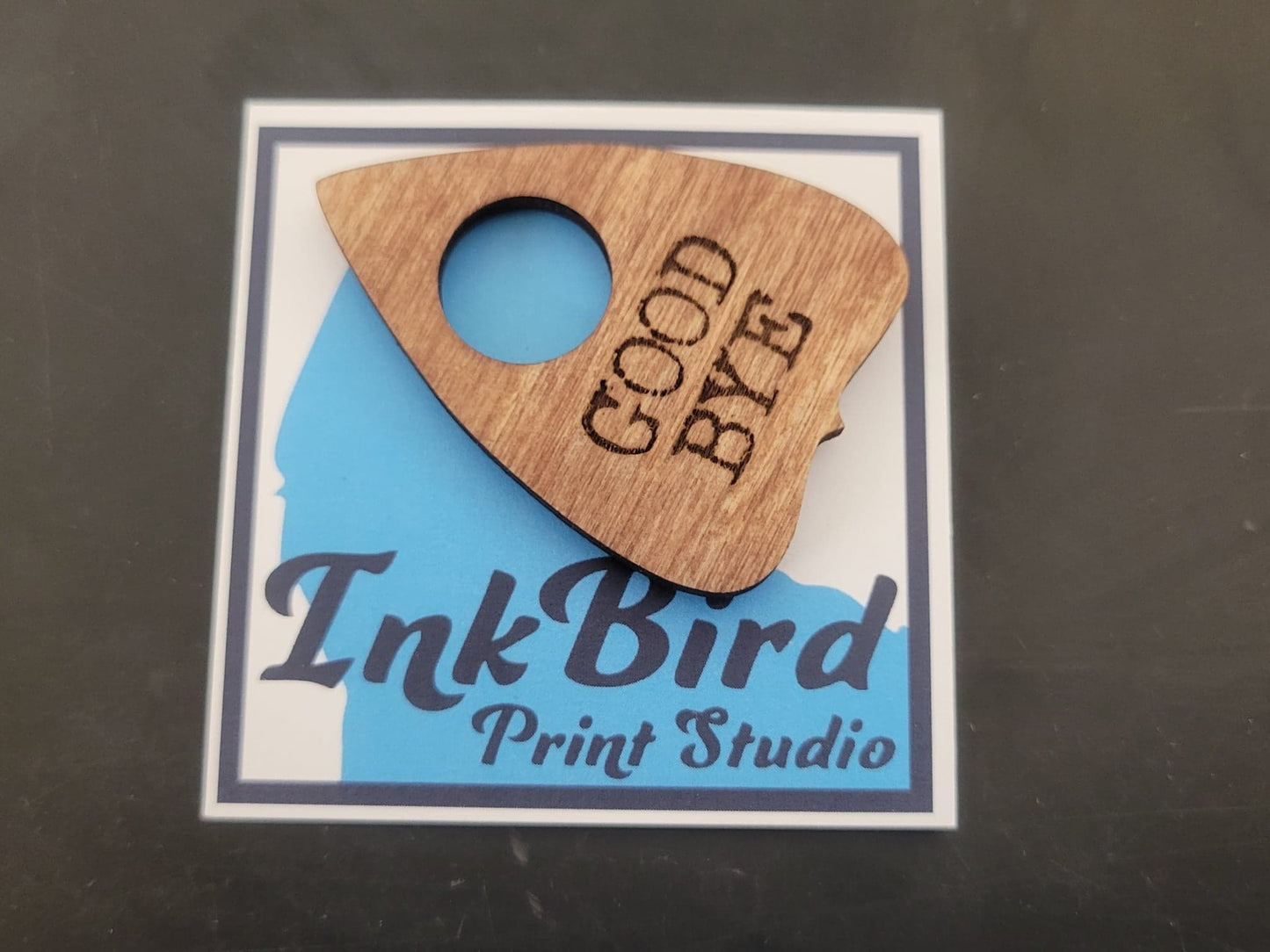 Laser-cut wooden Planchette Pin by InkBird Print Studio, available in large or small size, on card with blue and black studio logo.