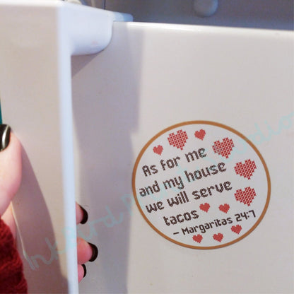 A sticker on a white surface, ideal as a Valentine's Day gift, showcases small red hearts and the witty text from the Cross Stitch Design Fridge Magnet by InkBird Print Studio LLC: "As for me and my house, we will serve tacos. Margaritas 24:7." A person's hand can be seen holding the edge of this playful fridge magnet.