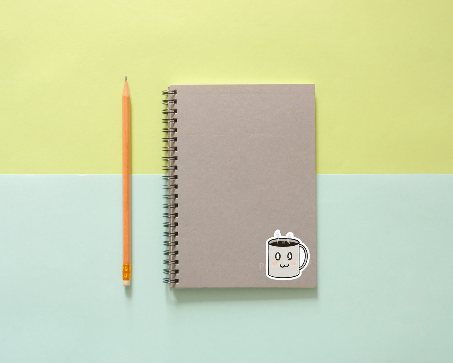 Gray spiral notebook decorated with a vinyl Adorable Coffee Cup Sticker, next to a yellow pencil on pastel yellow and blue backdrop.