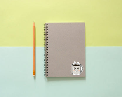 Gray spiral notebook decorated with a vinyl Adorable Coffee Cup Sticker, next to a yellow pencil on pastel yellow and blue backdrop.