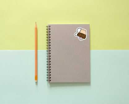Gray spiral notebook with a glossy vinyl Adorable Happy Tea Sticker by InkBird Print Studio, next to an orange pencil, on colorful background.